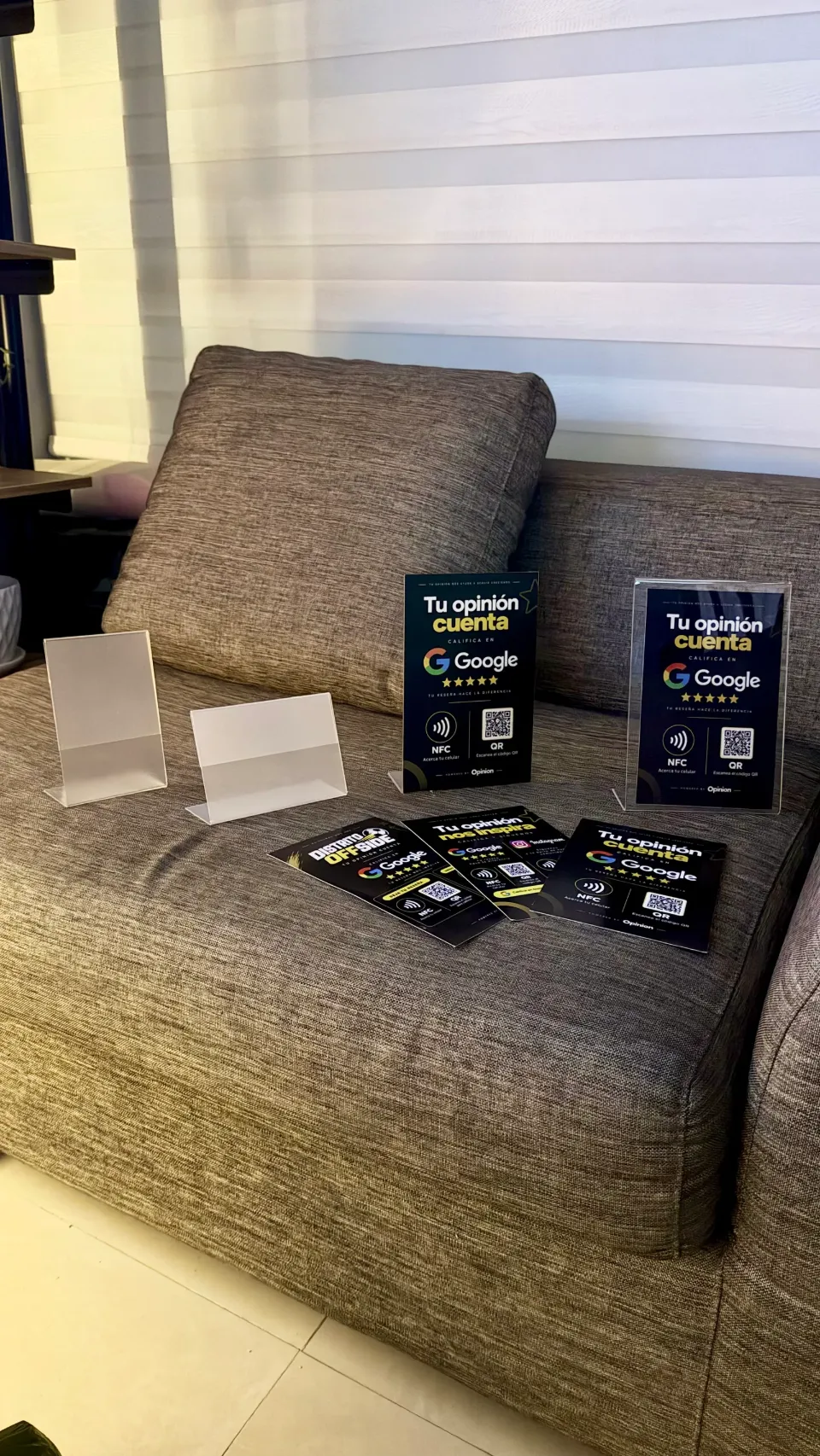Soportes reales de OPINION sobre un sofá: dos soportes transparentes vacíos, dos soportes negros con NFC y QR para reseñas de Google y tarjetas negras